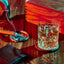 Glass of whiskey with ice on a wooden table, accompanied by a smoking cigar in an ashtray. A wooden box sits in the background, a leather wallet lies in the foreground.