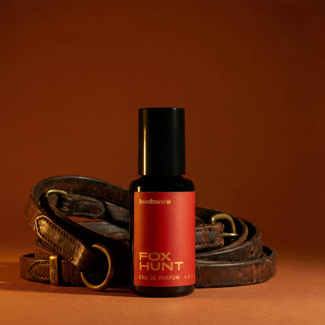A bottle of Beardbrand Fox Hunt Eau de Parfum stands in front of coiled brown leather straps on a warm brown studio background.
